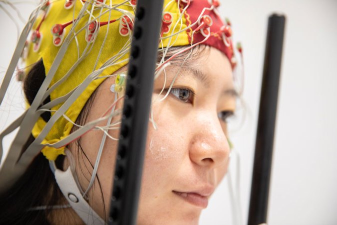 A study participant is facing to the right, wearing a yellow and red electroencephalogram (EEG) head gear that's tight around the participant's head like a swim cap. The head gear has electrodes attached to it, and it has a chip strap that goes around the bottom of the participant's face.